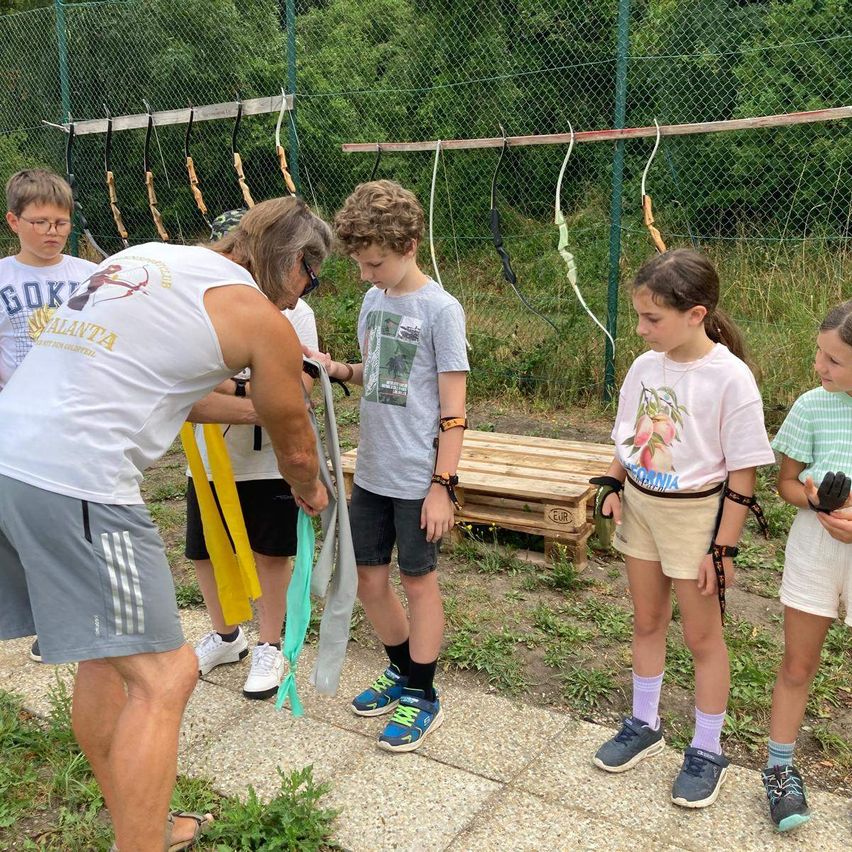 A group of children and an adult are standing in an outdoor area, possibly a park, with a wooden fence and a wooden structure in the background. The adult is holding a rope and appears to be teaching the children how to use it. The children are wearing bracelets and sneakers.