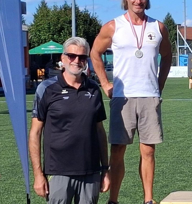 Zwei Männer stehen auf einem Feld, einer trägt eine Medaille und der andere ein schwarzes Shirt. Hinter ihnen ist ein grüner Schirm und ein Gebäude zu sehen.