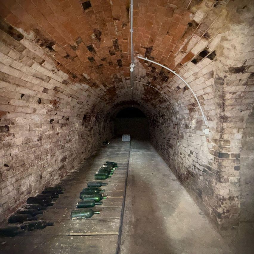 Ein gemauerter Tunnel mit auf dem Boden aufgereihten Flaschen, einer Überkopfleitung und einer weißen Kiste auf der rechten Seite.