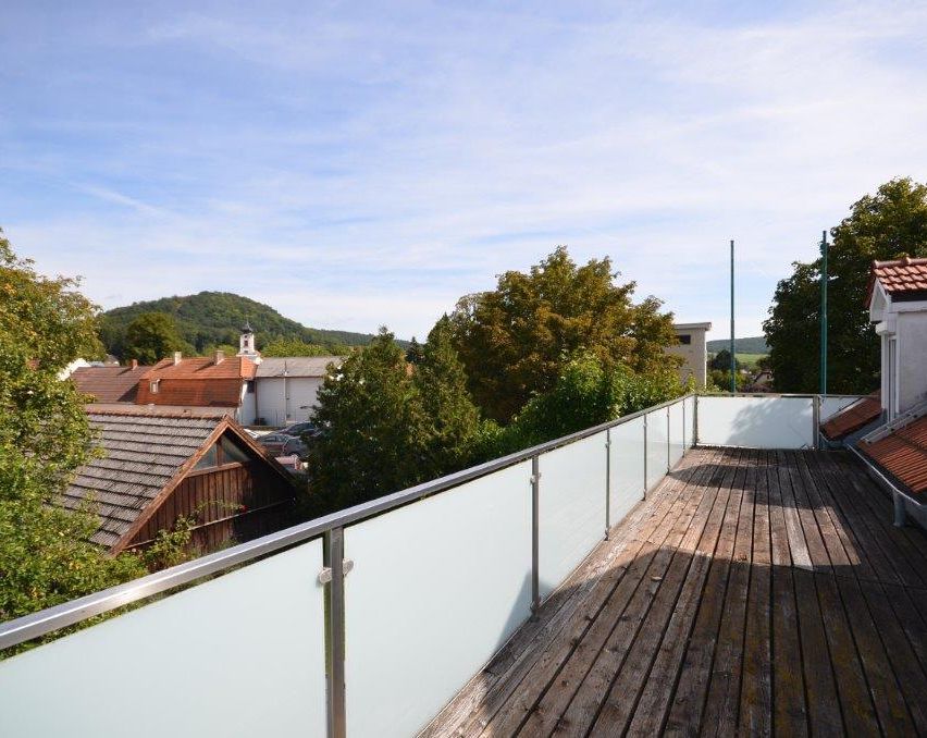 Ein Balkon mit Holzboden und Glasgeländern blickt auf eine Nachbarschaft mit Häusern und Bäumen unter einem klaren blauen Himmel.