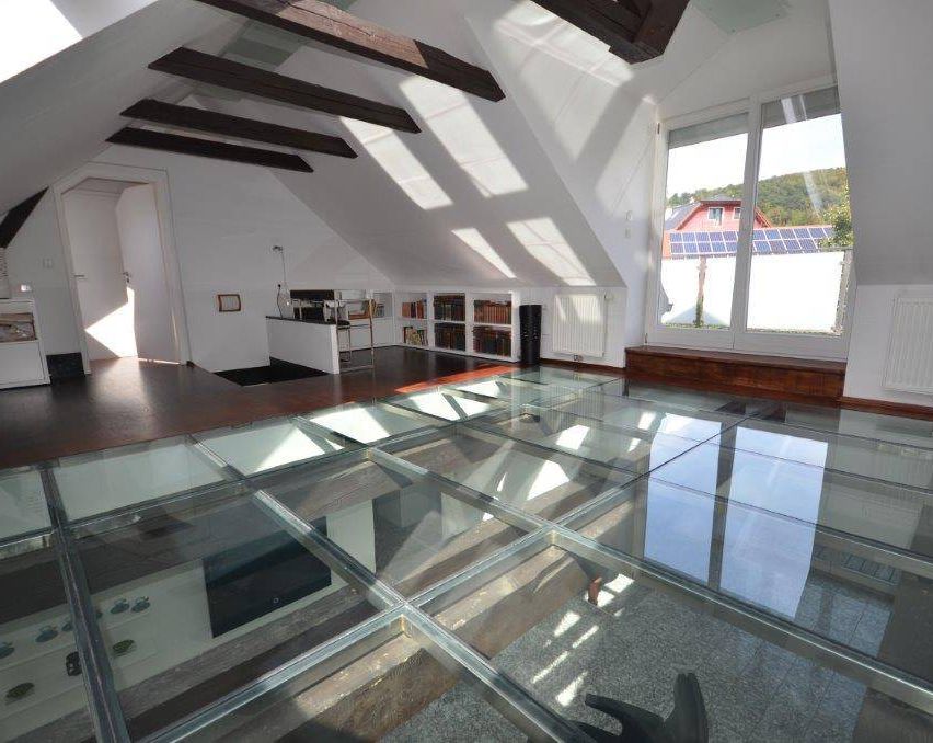Ein Dachboden mit Glasboden, der einen Raum mit weißer Tür, Bücherregalen, einem Schreibtisch und einem großen Fenster mit sichtbaren Solarpanelen draußen überblickt.