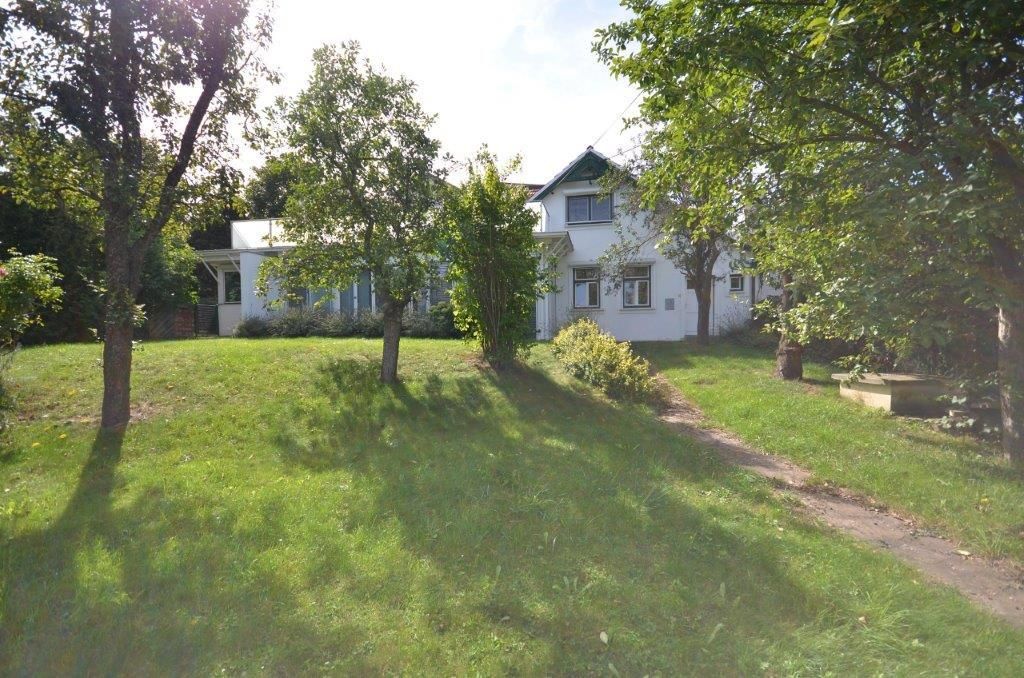 Ein weißes Haus mit einem grünen Dach steht auf einem großen, gepflegten Rasen mit verschiedenen Bäumen und Sträuchern. Der Rasen ist gut gepflegt und das Haus ist von Grün umgeben.