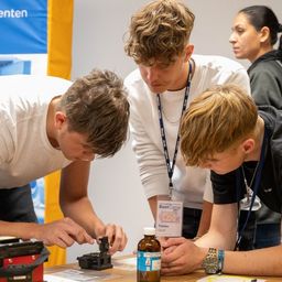 Drei Jungen in Laborkittel arbeiten an einem Projekt. Ein Junge hält eine Flasche, ein anderer justiert eine Maschine und der dritte beobachtet. Sie sind in einem Raum mit einem blauen Banner an der Wand.