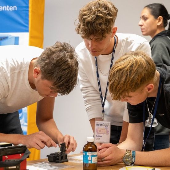 Drei Jungen in Laborkittel arbeiten an einem Projekt. Ein Junge hält eine Flasche, ein anderer justiert eine Maschine und der dritte beobachtet. Sie sind in einem Raum mit einem blauen Banner an der Wand.