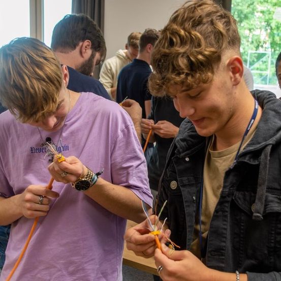 Zwei Männer sind mit einer Aufgabe beschäftigt, möglicherweise das Reparieren von Drähten. Einer trägt ein lila Hemd und der andere eine schwarze Jacke. Sie sind von anderen umgeben, mit einem Fenster im Hintergrund.
