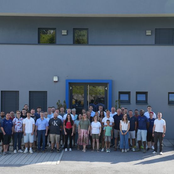 Eine Gruppe von Menschen steht vor einem modernen Gebäude mit Glasfenstern und einer blauen Eingangstür. Sie sind lässig gekleidet und scheinen für ein Foto zu posieren.