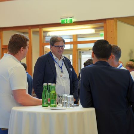 Mehrere Personen versammeln sich in einem Konferenzraum und unterhalten sich. Ein Mann mit Brille und Namensschild steht an einem Tisch mit grünen Flaschen und Gläsern. Ein anderer Mann in einem weißen Hemd schaut den Mann mit der Brille an.