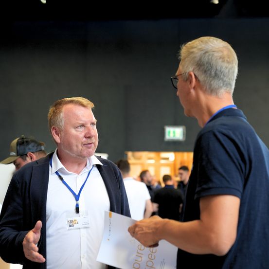 Zwei Männer unterhalten sich bei einer Veranstaltung. Einer hält ein Papier. Ein Notausgangsschild ist hinter ihnen.