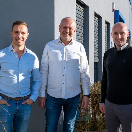 Drei Männer stehen vor einem Gebäude. Der Mann auf der linken Seite trägt ein hellblaues Hemd, Jeans und einen Gürtel. Der Mann in der Mitte trägt ein weißes Hemd und blaue Jeans. Der Mann auf der rechten Seite trägt ein schwarzes Hemd und graue Hosen.