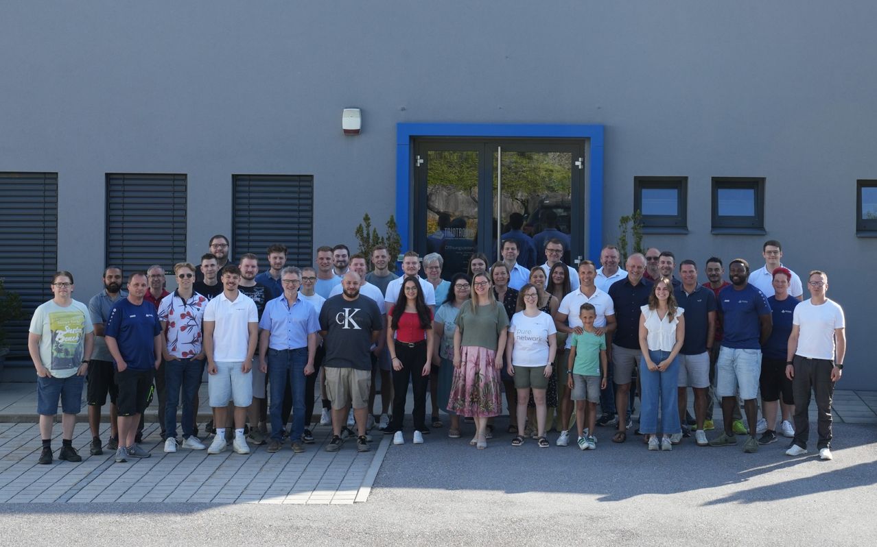 Eine Gruppe von Menschen steht vor einem Gebäude mit blauer Umrandung. Sie alle lächeln und scheinen für ein Foto zu posieren. Das Gebäude hat eine Glastür mit einem Spiegelbild von Menschen im Inneren.