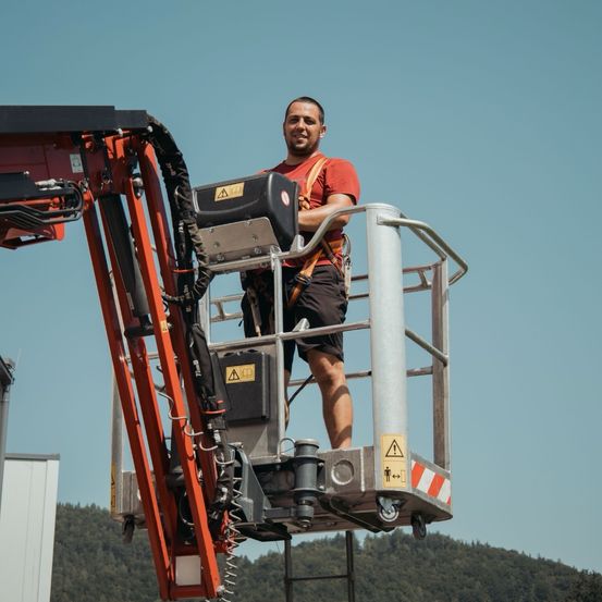 Ein Mann steht auf einem Scherenlift, trägt ein rotes T-Shirt und einen orangefarbenen Gurt und hält sich am Lift fest. Im Hintergrund sind Berge.