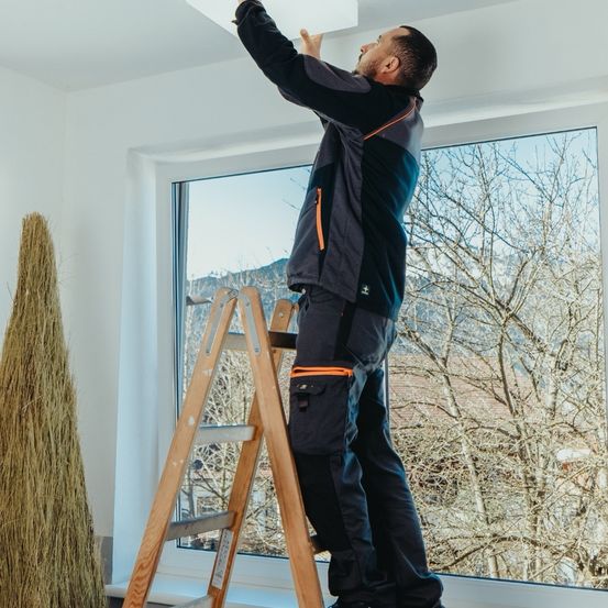 Ein Mann steht auf einer Holzleiter und repariert eine Deckenlampe in einem Raum mit weißen Wänden und einem Glasfenster, das Bäume draußen zeigt.