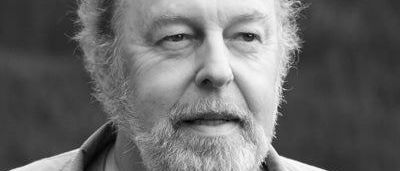 Schwarz-Weiß-Porträt eines älteren Mannes mit Bart und Schnurrbart, der eine helle Jacke trägt.