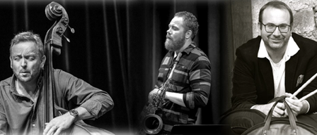 Drei Musiker in Schwarz-Weiß posieren für ein Foto. Der linke spielt Cello, der mittlere spielt Saxophon und der rechte hält Drumsticks.