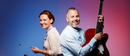 Ein Mann und eine Frau posieren für ein Foto, beide lächeln. Die Frau hält einen Taktstock und eine Zither, während der Mann eine Gitarre hält. Sie sitzen auf Hockern vor einem bunten Hintergrund.