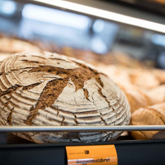 Ein rundes Brot mit rissiger, rustikaler Kruste steht auf einem Bäckereiregal, beschriftet mit 'Roggenmalzbrot'.