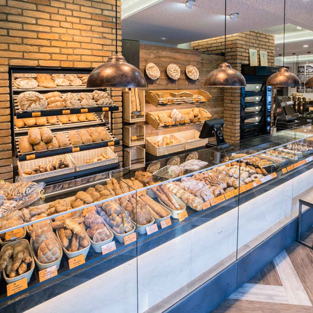 Eine Bäckerei-Vitrine mit verschiedenen Brotsorten, Croissants und Gebäck, mit Regalen voller Brot dahinter und kupfernen Pendelleuchten darüber.