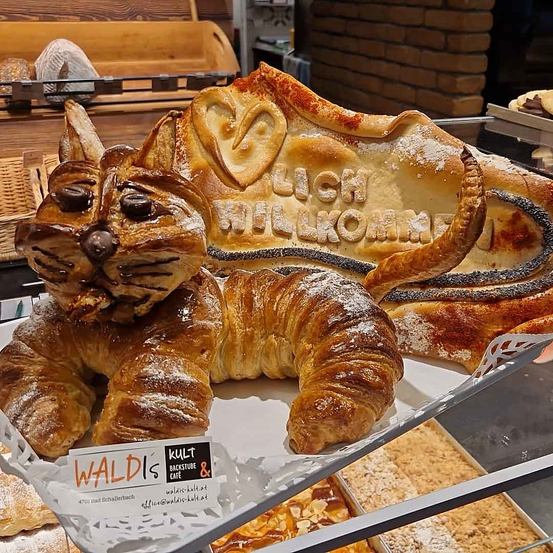 Eine katzenförmiges Gebäck aus Croissants mit Schokoladenchips als Augen und einem Schild darüber, das 'Lich willkommen' besagt.