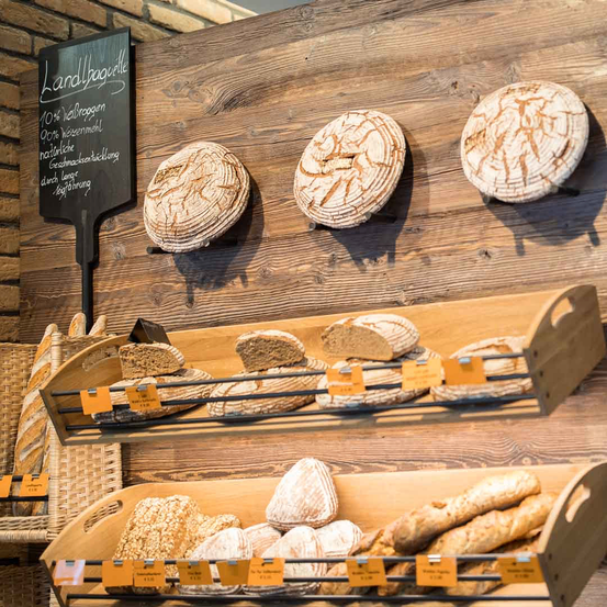 Eine hölzerne Ausstellung mit verschiedenen Brotsorten, einschließlich runder Laibe und Baguettes, auf Regalen angeordnet. Eine Tafel mit handgeschriebenem Text ist an der Wand befestigt.