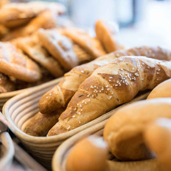 Nahaufnahme verschiedener Brotsorten, einschließlich Croissants mit Sesamkörnern, in Holzkörben auf einem Tisch angeordnet.