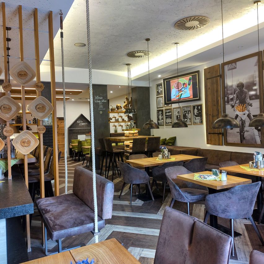 Ein Restaurant mit Holzmöbeln, hängenden Lampen und Wanddekorationen. Tische und Stühle sind angeordnet, und einige Tische haben gelbe Tischsets. Es gibt eine Theke mit einer Tafel und eine Tafel an der Wand.