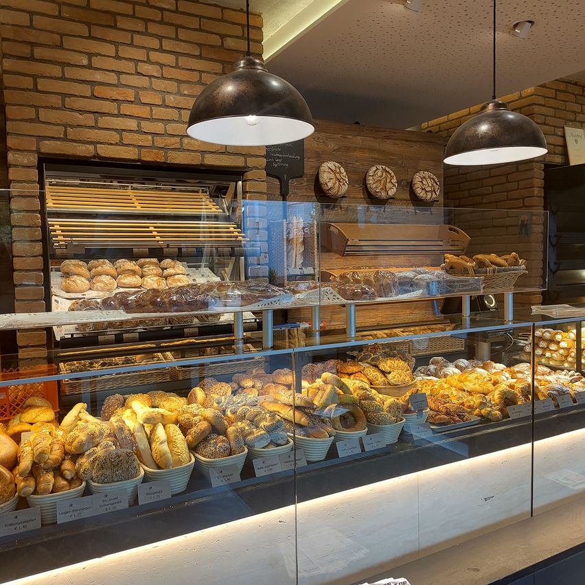 Ein Bäckerei-Schaufenster mit einer Auswahl an Brot und Gebäck. Die Regale sind gefüllt mit verschiedenen Brotsorten, darunter Vollkorn- und Sauerteigbrot. Die Gebäckauswahl umfasst Croissants, Dänisch und andere süße Leckereien. Das Schaufenster besteht aus Glas und hat eine Ziegelsteinwand dahinter.