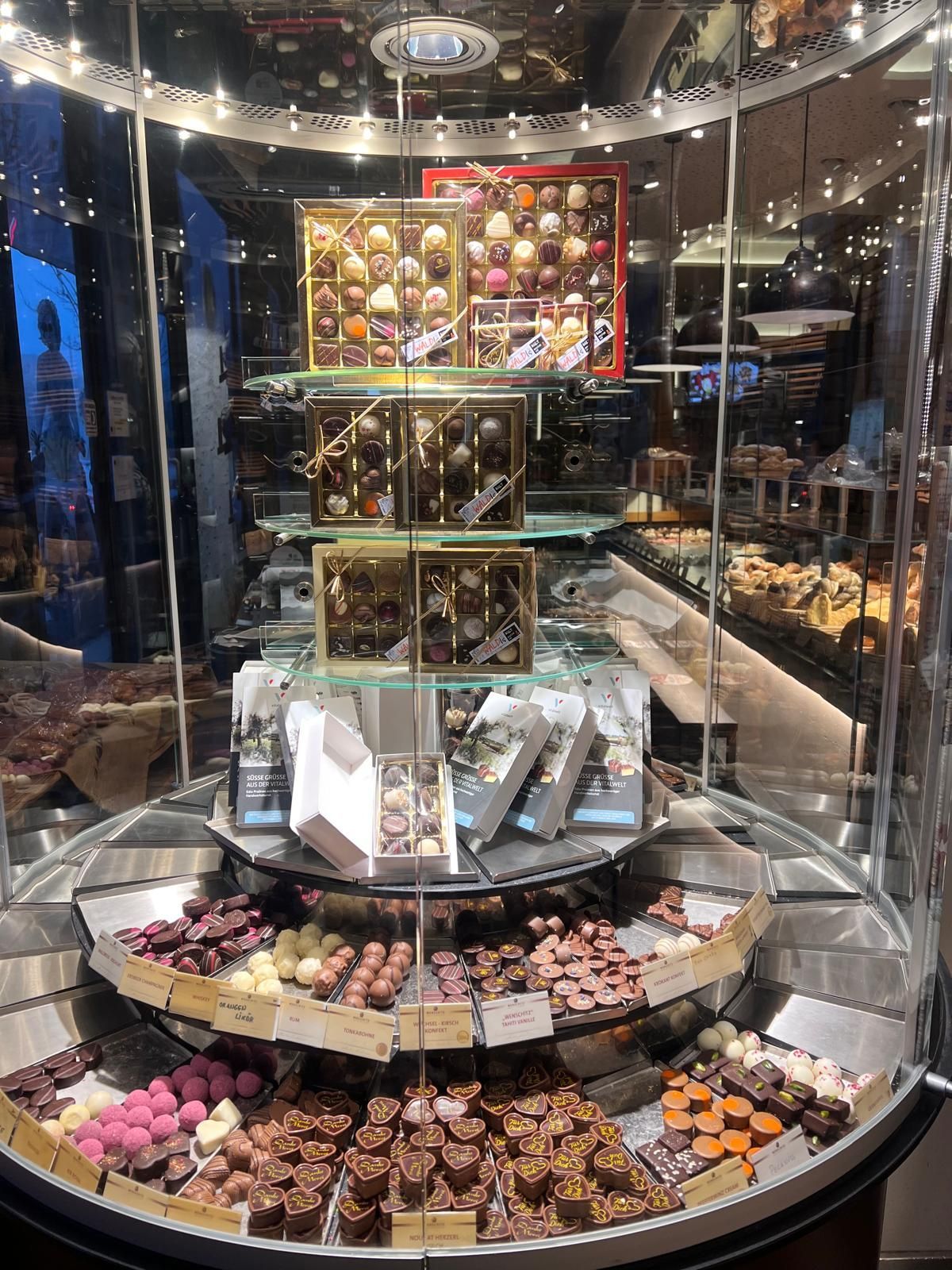 Ein Glasvitrine präsentiert eine mehrstöckige Anordnung von verschiedenen Schokoladen. Jede Ebene ist mit Schokoladen in dekorativen Schachteln gefüllt, umgeben von anderen Süßigkeiten.