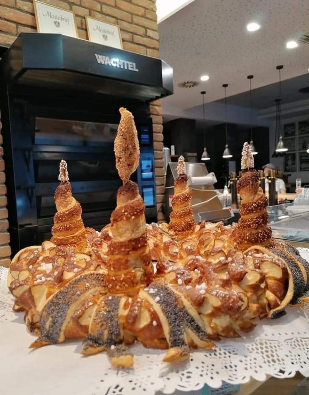 Eine Ausstellung von kunstvoll gestalteten Gebäckstücken auf einem weißen Tischtuch vor einer Küche mit einer Ziegelsteinwand und einem schwarzen Ofen.