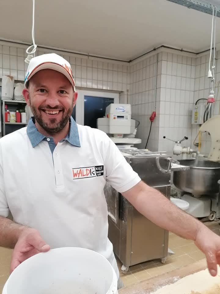 Ein Mann, der ein weißes Hemd mit dem Aufdruck WALDIS trägt, lächelt und hält einen weißen Teller in einer Küche.