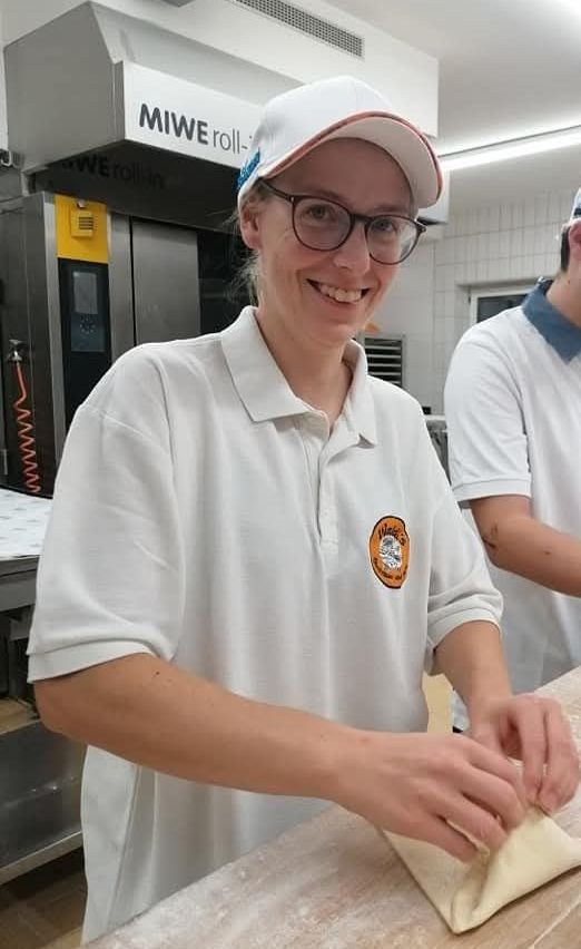Eine lächelnde Frau mit Kappe und Brille steht in einer Küche und knetet Teig. Hinter ihr steht ein Mann, der ebenfalls in der Küche arbeitet. Über ihnen ist ein Ofen mit einem Markennamen zu sehen.