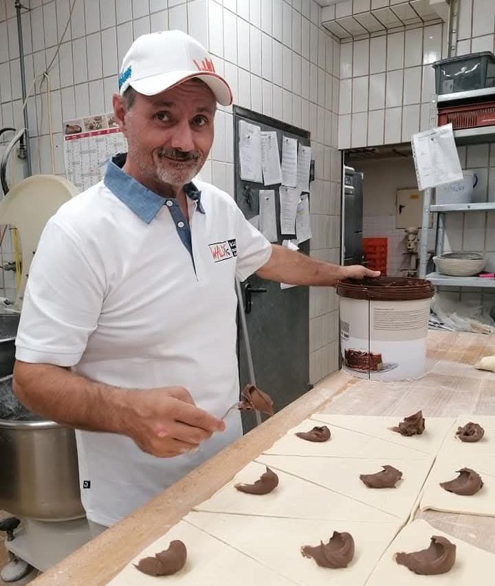 Ein Mann in einer Küche, der ein weißes Hemd und eine Kappe trägt, arbeitet mit Schokolade auf einem Holztisch. Er hält eine Palette und hat schokoladeüberzogene Gebäckstücke auf dem Tisch angeordnet. Hinter ihm befinden sich Regale mit Aufbewahrungsboxen, einem Eimer und angehefteten Papieren an der Wand.