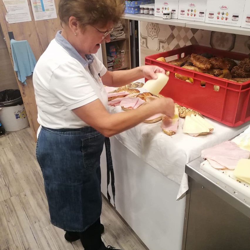 Eine Frau bereitet Sandwiches an einer Theke in einer Bäckerei zu. Sie trägt ein weißes Hemd und eine Jeansschürze. Es gibt Brot, Schinken und Käse auf dem Tresen.