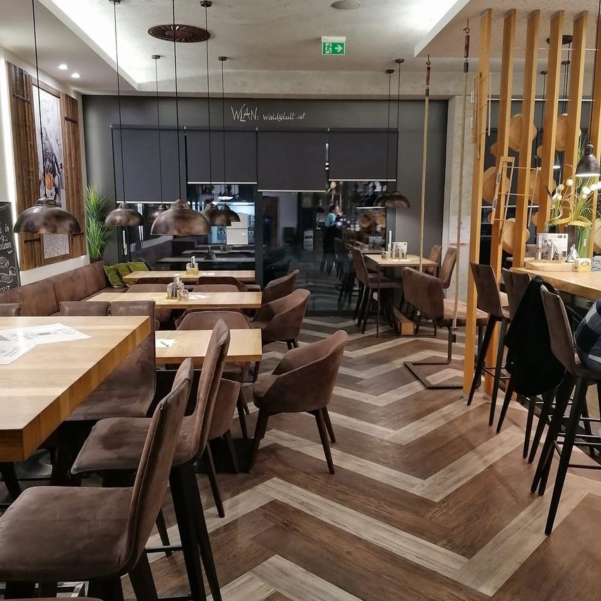 Ein Restaurant mit modernem Innendesign verfügt über Holzböden und braune Stühle. Es gibt Tische mit verschiedenen Gegenständen, und über ihnen hängen Lampen. Eine Person steht in der Nähe einer Glastür, und eine Tafel ist an der Wand montiert.