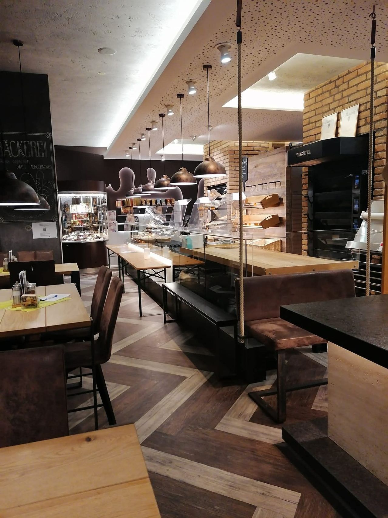 Eine moderne Bäckerei mit Ziegelwänden, einer langen Theke und Tischen und Stühlen für Kunden. Es gibt hängende Lampen und Regale mit verschiedenen Artikeln.