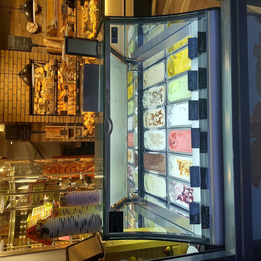 Eine Eistheke mit mehreren Geschmacksrichtungen auf Tabletts, in einer Bäckerei mit Ziegelwänden und verschiedenen Backwaren.