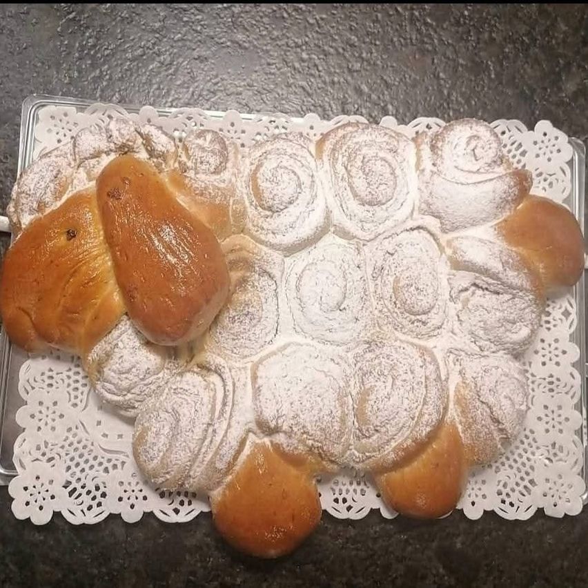 Ein wie ein Schaf geformter Teig mit Puderzucker darauf liegt auf einem weißen Spitzentuch. Der Teig hat eine goldbraune Farbe und eine leicht glänzende Erscheinung.