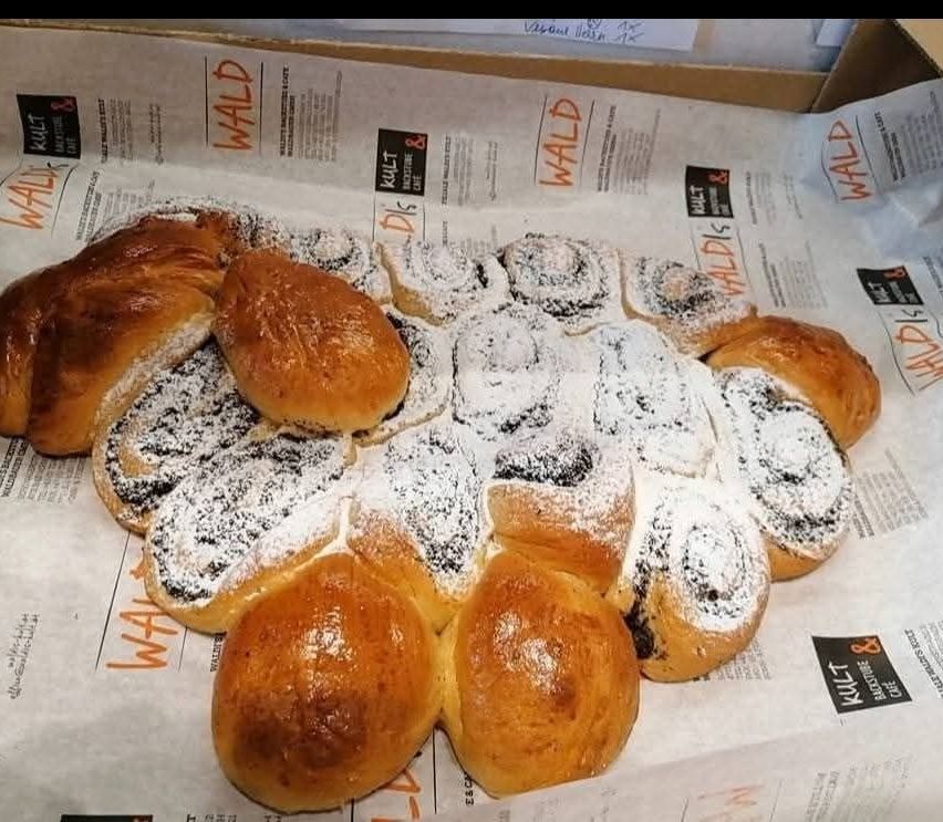 Ein Nahaufnahme von einer Sammlung von Gebäck, die in Form einer Blume angeordnet sind, jedes mit Puderzucker bestreut. Die Gebäckstücke scheinen Brötchen zu sein, einige mit sichtbaren Füllungen. Sie sind auf einem Papier mit Text platziert.