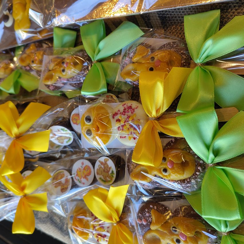 Mehrere Packungen Ostersüßigkeiten mit gelben und grünen Schleifen. Jede Packung enthält ein Schokoladen-überzogenes Leckerli, einige mit einem kleinen Gesichtsdesign. Die Leckerlis sind zur Präsentation in Plastik eingeschweißt.