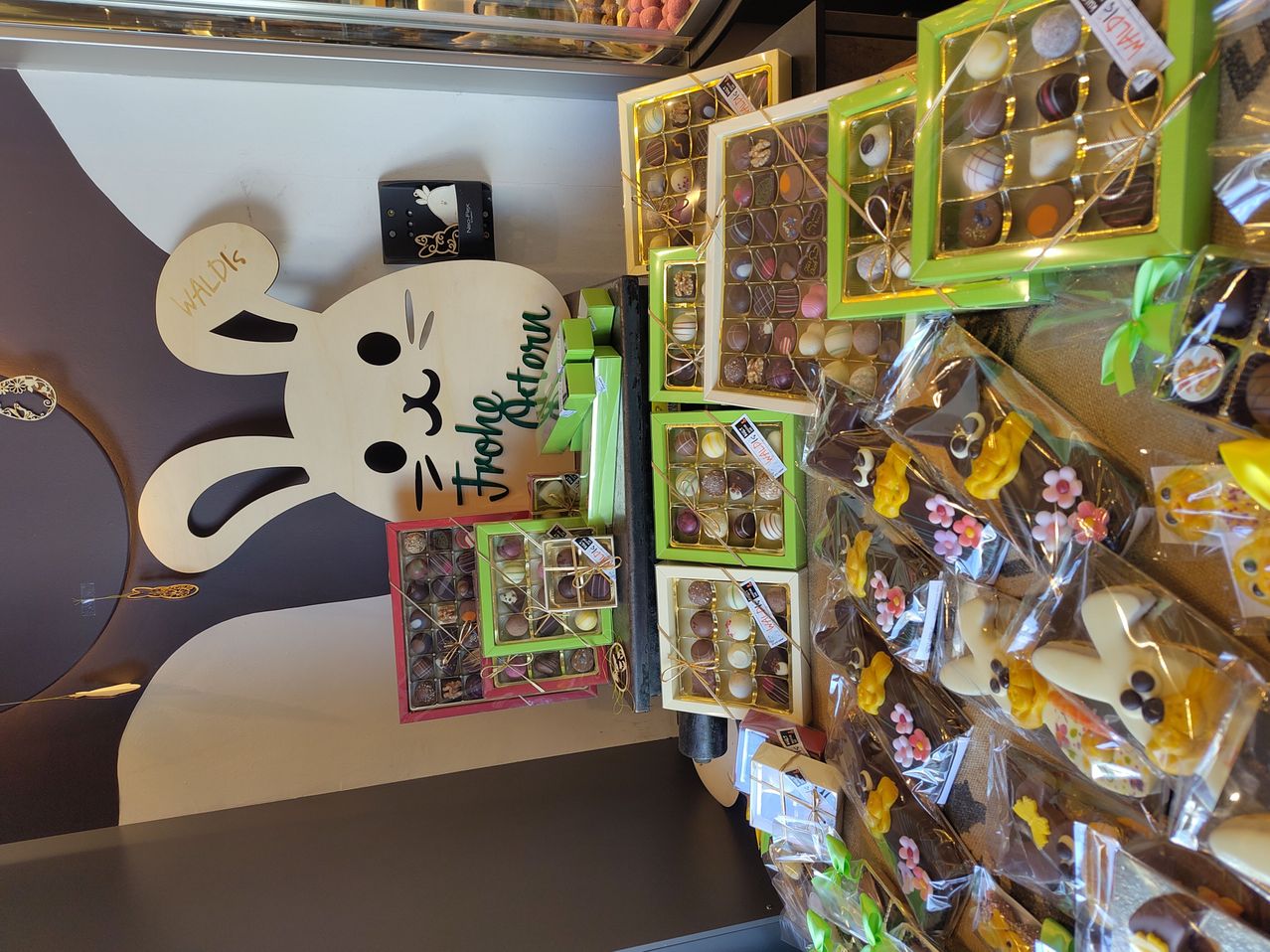 Eine Ausstellung von verschiedenen Schokoladen und Süßwaren, einschließlich eines großen Schildes in Form eines Hasen, mit dem Text 'Frohe Ostern' darauf geschrieben. Die Schokoladen sind ordentlich in verschiedenen Geschenkboxen angeordnet und in Plastik eingewickelt.