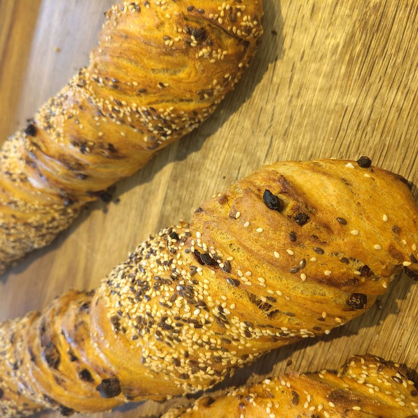 Nahaufnahme von zwei spiralförmigen Brotlaiben mit Sesamkörnern auf einer Holzoberfläche. Ein Laib hat sichtbare dunkle Rosinen.