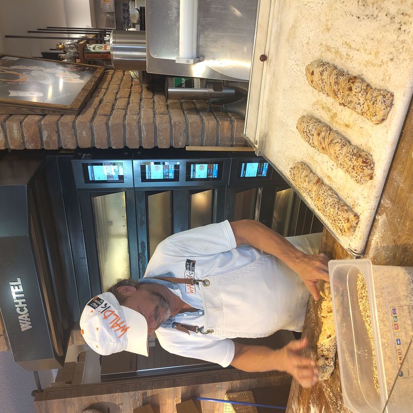 Ein Mann in einer Bäckerei, der eine weiße Schürze und einen Hut trägt, beugt sich über einen Tisch mit einem Tablett Brot. Der Tisch ist aus Holz und hinter ihm befindet sich eine Ziegelmauer.