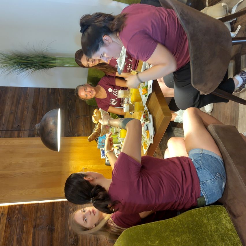 Vier Frauen in weinroten Shirts und Jeansshorts sitzen um einen Holztisch herum, bereiten Essen und Getränke zu. Der Tisch ist mit verschiedenen Gegenständen bestückt, und eine Lampe steht links.