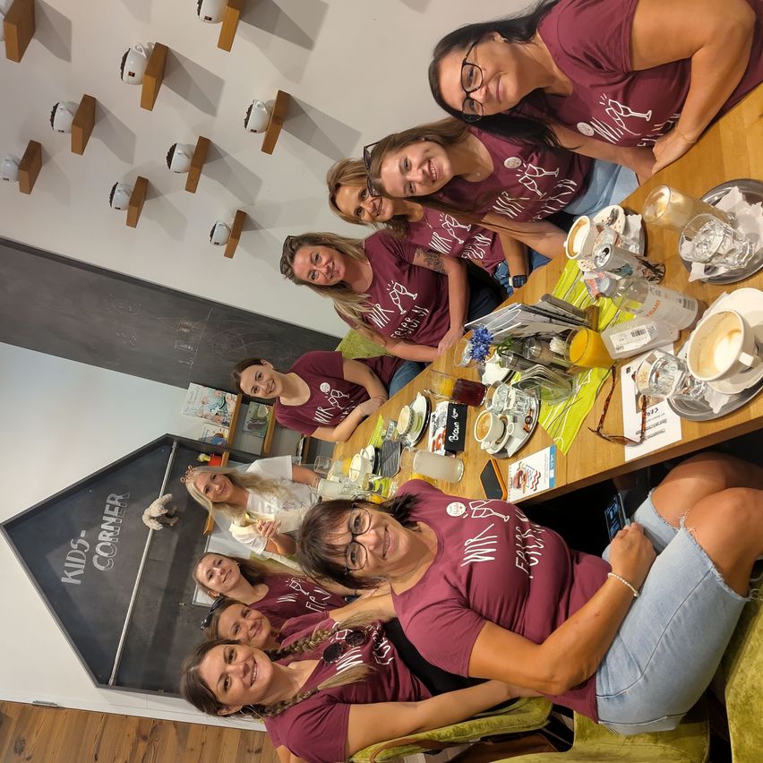 Eine Gruppe von Frauen in passenden Weinroten Shirts lächelt und posiert für ein Foto an einem Tisch mit Getränken und Essen.