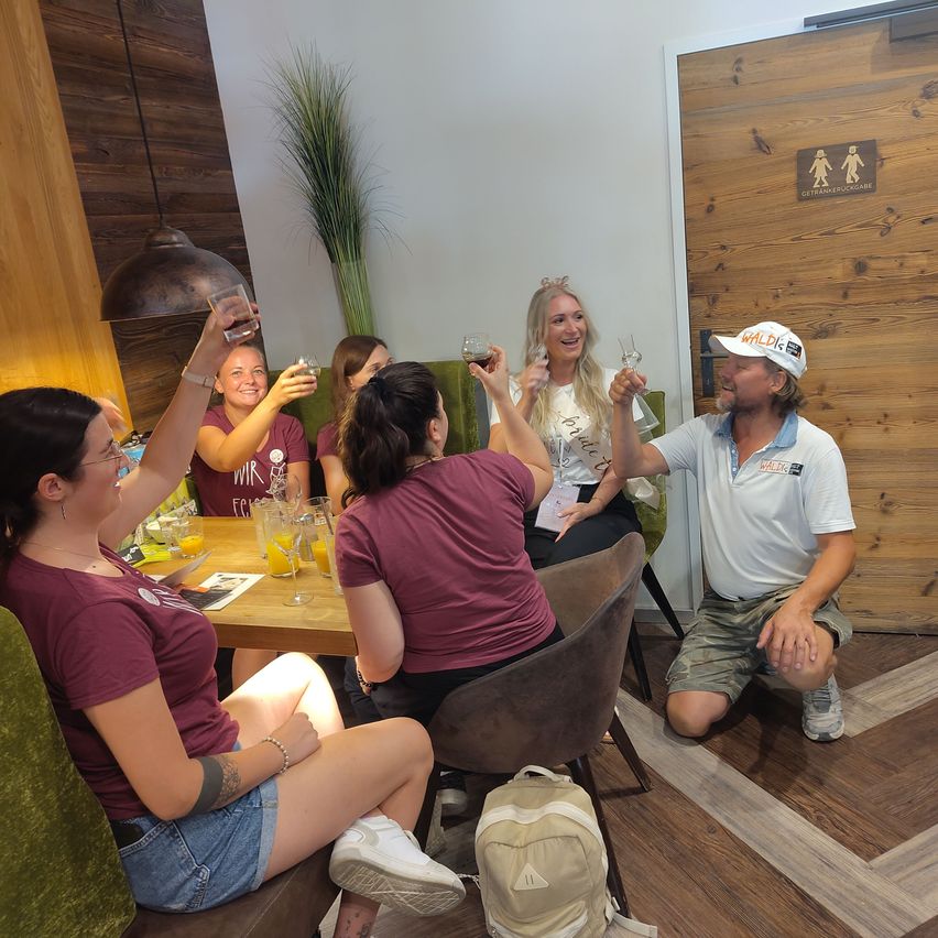 Eine Gruppe von Menschen sitzt in einem Restaurant um einen Tisch herum und hebt ihre Gläser zum Anstoßen. Ein Mann in einem weißen Hemd kniet vor ihnen.