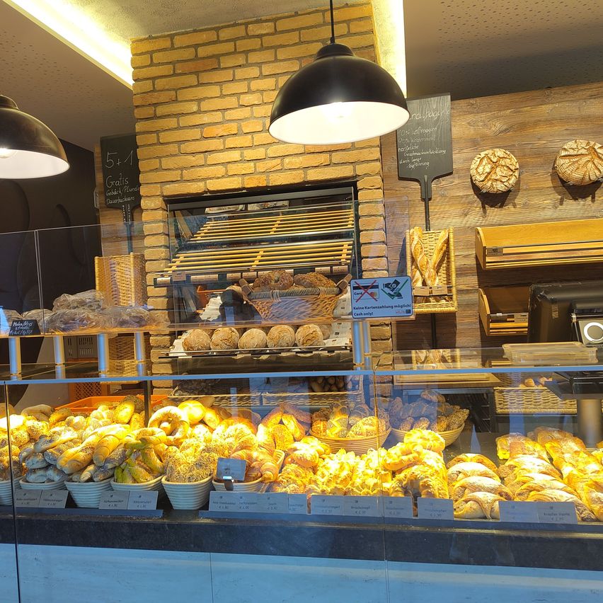 Eine Bäckerei-Auslage mit verschiedenen Broten und Gebäck unter einer Lampe. Regale mit Broten sind an der Wand, und es gibt ein Schild, das Kartenzahlung verbietet.