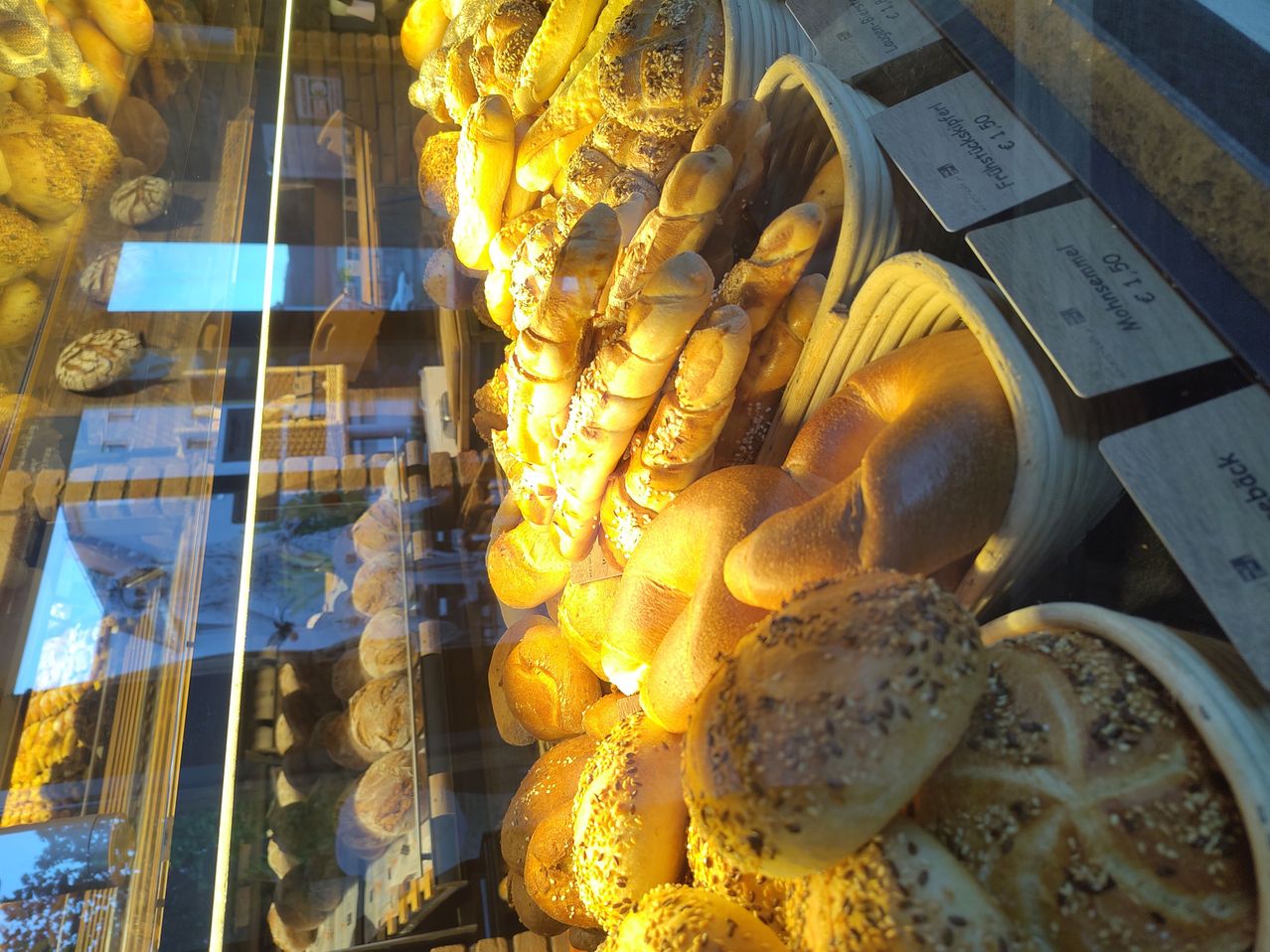 Eine Vitrine gefüllt mit verschiedenen Arten von Brot, einschließlich Brötchen, Laibe und Brötchen, jeweils mit einer anderen Kruste. Das Licht reflektiert auf der Glasvitrine und hebt die goldene Farbe des Brotes hervor.