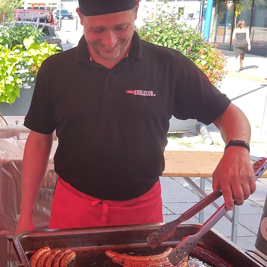 A man in a black chef hat and red apron is grilling sausages on a grill outdoors.