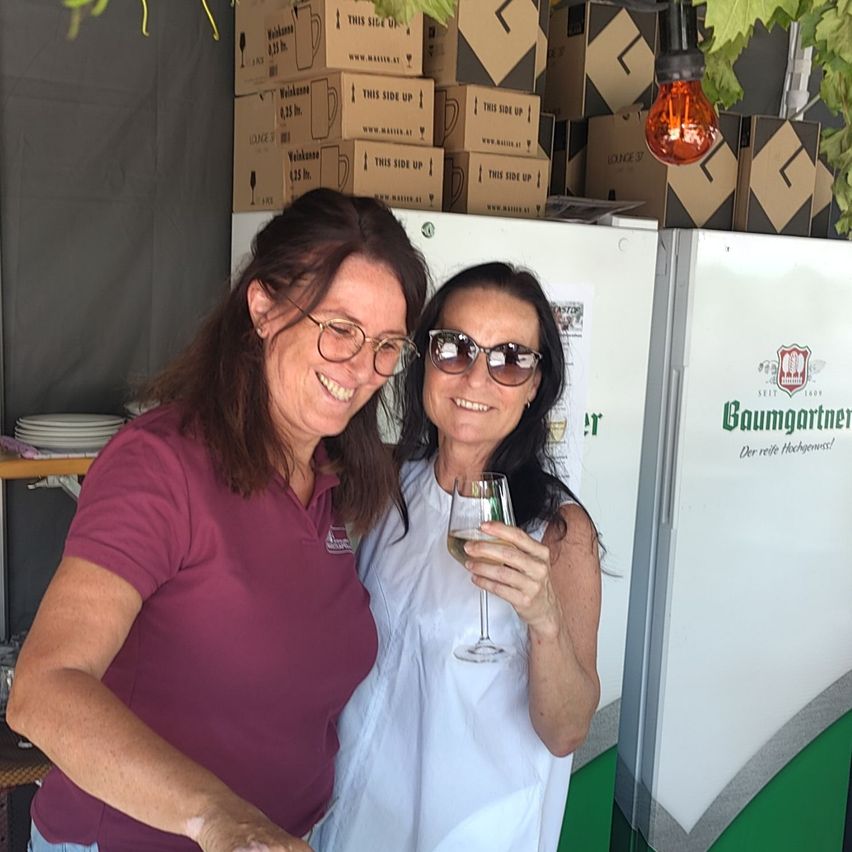 Two women smiling and posing for a photo, one holding a wine glass, standing in front of a refrigerator with a logo and boxes of wine stacked behind them.