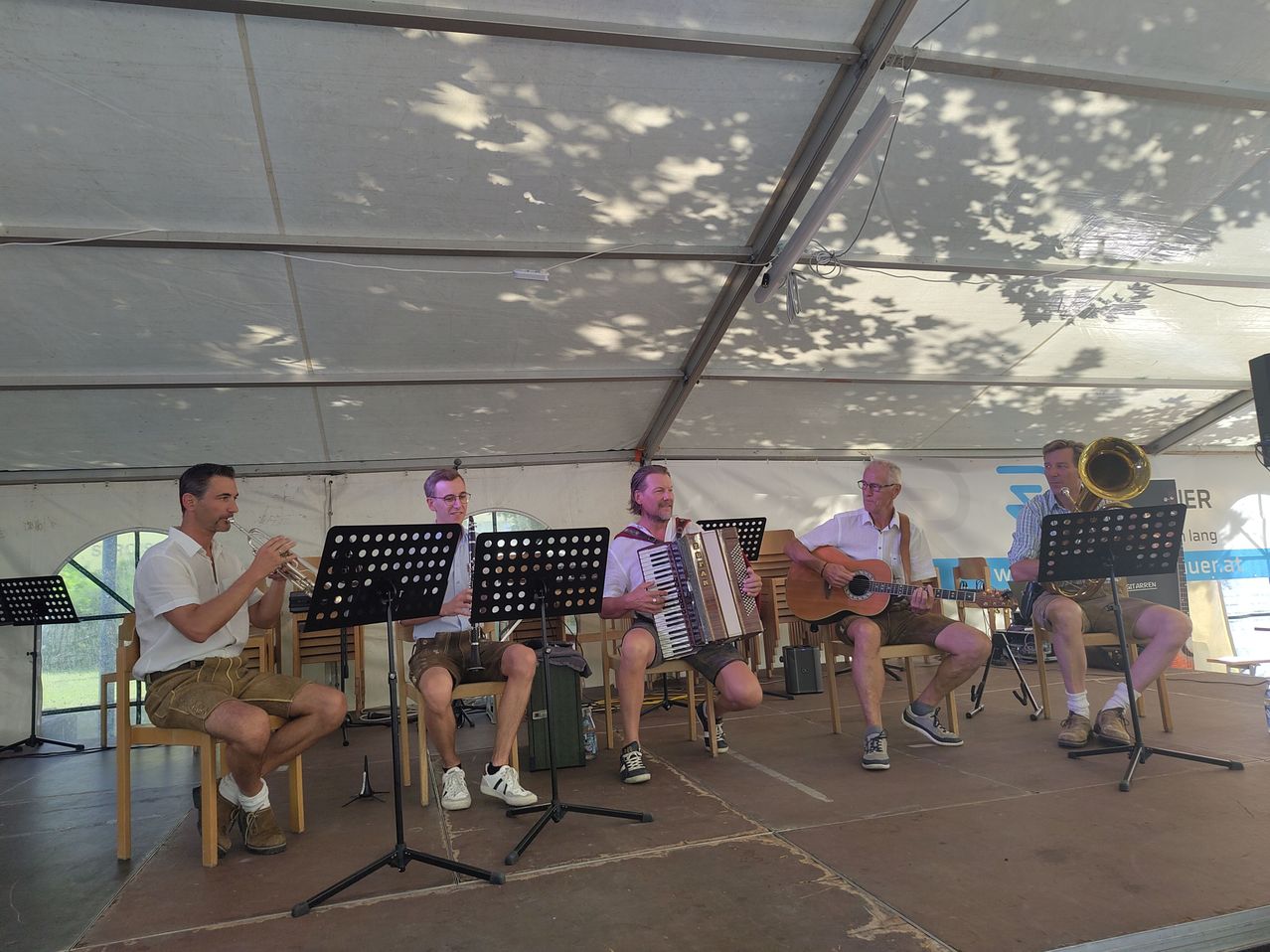 Five men in traditional attire are performing on stage, with one playing the trumpet, another the accordion, and others playing guitar and trombone.