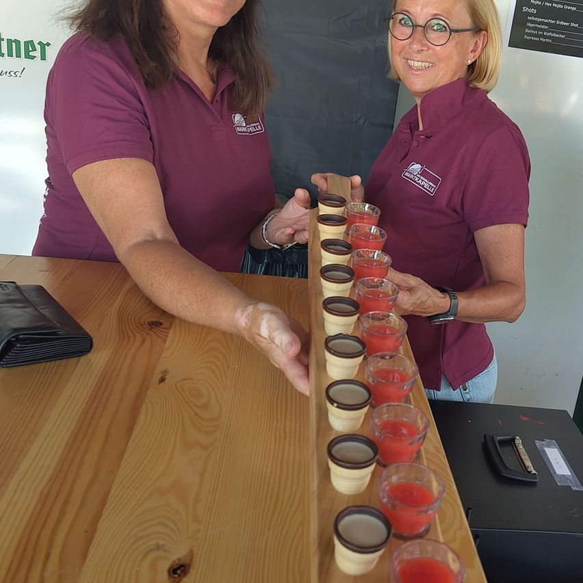 Two women in purple shirts are standing behind a wooden table, smiling and holding a tray of red-colored drinks in small cups.